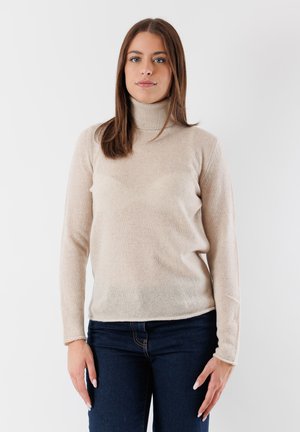 Giovane donna con capelli castani lisci che indossa un maglione beige a collo alto e jeans blu scuro, in piedi contro uno sfondo bianco semplice.