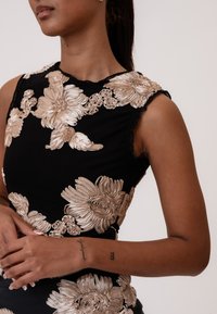 Sort kjole med indviklet beige blomsterbroderi, teksturerede detaljer og bølgede ærmekanter, der fremhæver en tætsiddende silhuet.