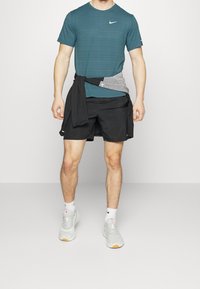 Man wearing teal short-sleeve Nike shirt, black shorts, white socks, white sneakers, with black and gray jacket tied around waist.