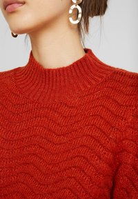 Červený pletený svetr s texturou, vysokým žebrovaným výstřihem a vlnitým vzorem. Detail ukazuje část límce a náušnice.