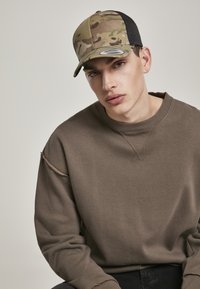Vrachtwagenchauffeur pet met een camouflage ontwerp en een zwarte gaas achterkant. Model draagt een bruine sweatshirt met een gestikt detail op de mouw.