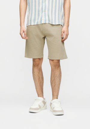 Mann trägt beige knieumspielte Shorts, weiße und beige Sneaker sowie ein hellfarbenes, gestreiftes Kurzarmhemd und steht vor weißem Hintergrund.