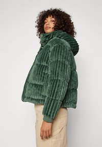 Groene corduroy pufferjack met een capuchon, met ribbeltextuur, ritssluiting en elastische boorden. Zijkant toont het dikke ontwerp.