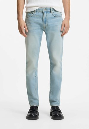 Mann trägt hellblaue Slim-Fit-Jeans, schwarze Leder-Slipper und ein weißes, strukturiertes Kurzarmoberteil, steht vor weißem Hintergrund.