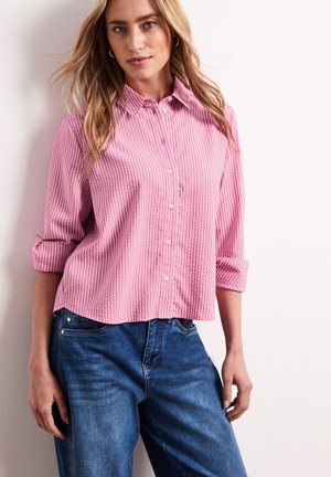 Femme portant une chemise à rayures verticales rose et blanc à boutons avec les manches retroussées et un jean bleu, debout contre un mur blanc.