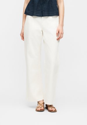 Žena oblečená v bielych širokých nohaviciach, hnedých sandáloch na remienky a tmavomodrom tielkovom topu s gombíkmi, stojaca pred jednoliatym pozadím.