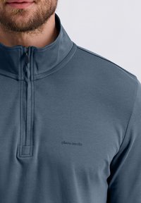 Uomo che indossa un pullover blu a zip quarti con il discreto logo "pierre cardin" sul petto, mostrando la parte bassa del volto e la barba su uno sfondo semplice.