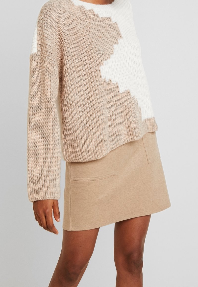 Pull en maille beige avec motif zigzag blanc sur le devant ; assorti à une mini-jupe beige avec poches latérales.