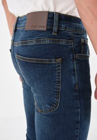 Jeans de mezclilla azul oscuro con corte ajustado, que cuentan con una etiqueta de cuero marrón con "PIER•ONE", costuras en contraste y múltiples bolsillos.