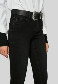 Black denim jeans with slight sheen, featuring a fitted design and visible pockets. Paired with a black leather belt and silver buckle.