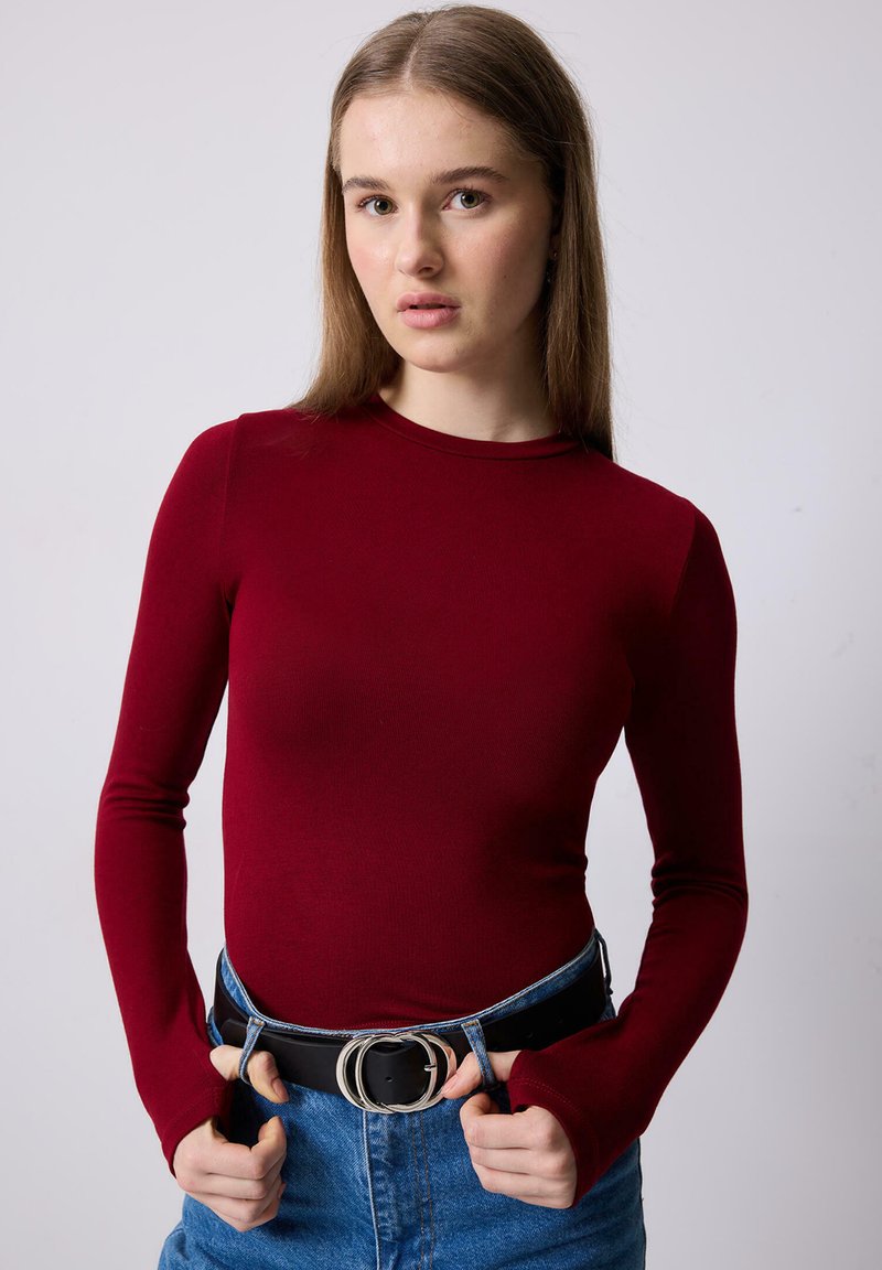 Νεαρή γυναίκα με ίσια ανοιχτό καστανά μαλλιά που φοράει εφαρμοστή μπλούζα μακριάς φανέλας σε μπορντό χρώμα και μπλε τζιν με μαύρη ζώνη διπλού δακτυλίου.