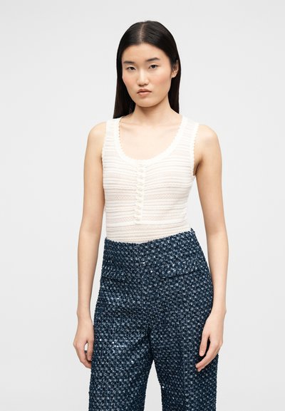 Γυναίκα με μακριά σκούρα μαλλιά, που φοράει ένα αμάνικο λευκό υφασμάτινο τοπ και ψηλόμεσα σκούρα μπλε παντελόνια με σχέδια, στέκεται σε απλό φόντο.