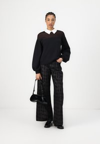 Schwarzer Pullover mit durchsichtiger Oberseite, weißem Kragen; gemusterte schwarze weit geschnittene Hose; kleine schwarze Handtasche; schwarze Schuhe mit Schnürdetails.