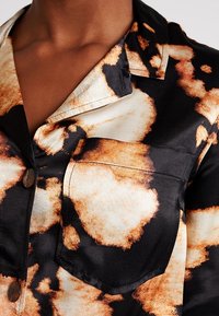 Satin skjorta med svart bas och abstrakta beige och orange fläckar, en kraag med urtag och knappstängning. Inkluderar en bröstficka.