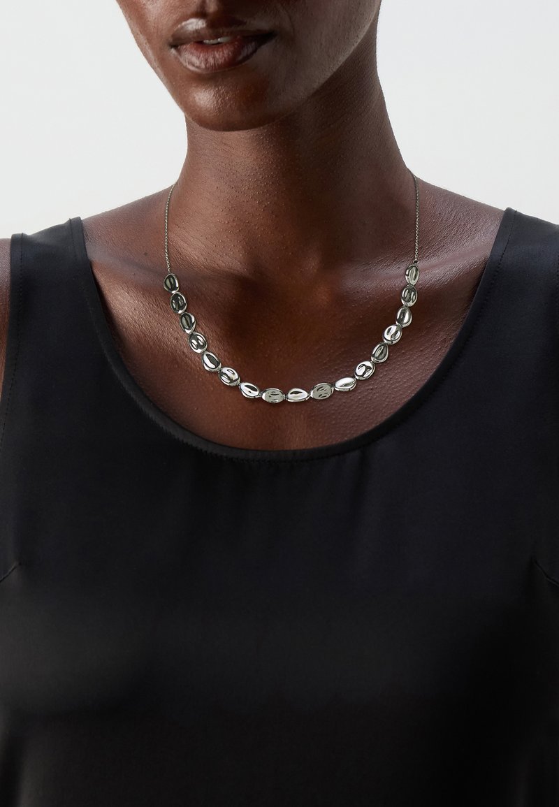 Stříbrně tónovaný náhrdelník s oválnými korálky s lesklým povrchem, nošený přes černý top. Zobrazuje jemný design řetízku.