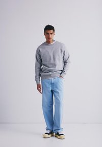 Pull gris à col rond avec des manches à bord-côte, associé à un jean droit bleu clair et des baskets noires avec des accents crème, se tenant devant un fond blanc.