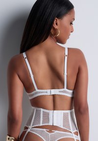 Ensemble de lingerie en dentelle blanche comprenant un corset structuré avec des bretelles réglables, une fermeture à crochets et œillets dans le dos, et des détails en motif géométrique.