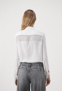 Blusa bianca con dettagli di frange sul retro e accenti argentati. Maniche lunghe con frange ai polsini. Indossata con pantaloni grigi a vita alta.