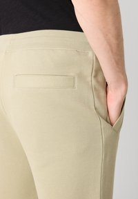 Light beige sweatpants with a ribbed waistband. Features a side pocket and a stitched fabric patch pocket on the back. Smooth texture.