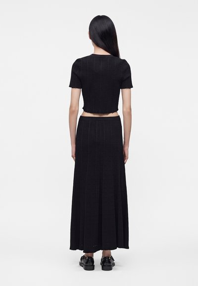 Femme aux longs cheveux noirs portant un crop top noir à manches courtes et une longue jupe, debout de dos sur un fond blanc uni.