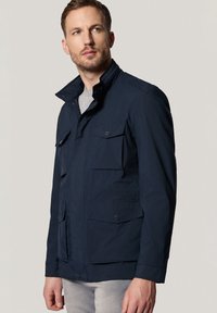 Granatowa, lekka kurtka z wieloma przednimi kieszeniami, zapięciem na guziki przy kołnierzu oraz gładką teksturą, charakteryzująca się opływowym wzornictwem.