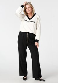 Witte top met lange mouwen, een zwarte V-hals en de tekst "BISOU PARIS", gecombineerd met zwarte broek met wijde pijpen en zwarte schoenen met studs.