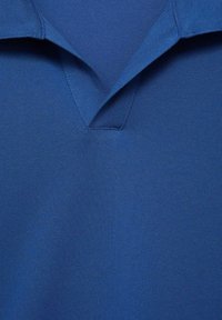 Pikeepaita tasaisessa sinisessä, jossa on teksturoitu kangas, V-pääntiellä ja lyhyillä hihoilla. Sauman yksityiskohdat näkyvät designissa.