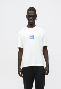 Biele bavlnené tričko s modrým obdĺ�žnikovým logom "HUGO". Nosené s čiernymi nohavicami s čiernym potlačeným nápisom "HUGO". Jednoduchý dizajn s okrúhlym výstrihom.