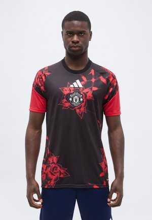 Młody mężczyzna ubrany w czarno-czerwoną koszulkę piłkarską Manchester United z czerwonymi wzorami róż i logo Adidas, stojący na jednolitym białym tle.