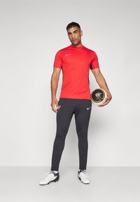 Rød Nike kortærmet top med sorte detaljer, sorte tætsiddende bukser og hvide fodboldstøvler. Model holder en fodbold.