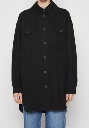 Sort oversize skjortejakke med knapper og to klaplommer på brystet, båret over blå jeans, vist fra nakke til midt på låret.