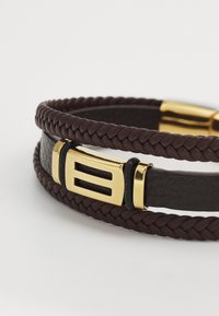 Brățară din piele împletită de culoare maro, având un închizător de culoare aurie cu un design rectangular și accente negre. Textură netedă pe toată suprafața.