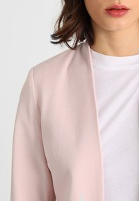 Světlá růžová sako s hladkým povrchem a otevřeným předním designem, s jemně zakřiveným límcem a dlouhými rukávy. Pod ním je vidět bílá tričko.