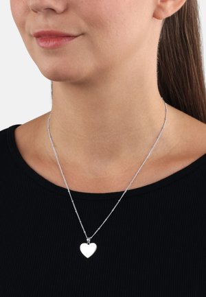 Collana con pendente a forma di cuore in argento, realizzata con una delicata catena, caratterizzata da anelli lisci alternati. Indossata su un top nero a coste con scollatura rotonda.