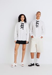 To modeller står ved siden av hverandre. De har på seg hvite skjorter med svarte bånd-lignende detaljer. Kvinnen har på seg svart skjørt; mannen har på seg beige shorts.