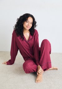Burgundy satinpyjamas med randig textur, långärmad skjorta med knappar och löst sittande byxor, med marinblå accenter.