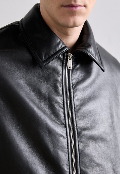 Veste en cuir noir avec un grand col et une fermeture éclair en argent. Texture lisse avec des détails de grain subtils. Vue en gros plan de la poitrine vers le haut.