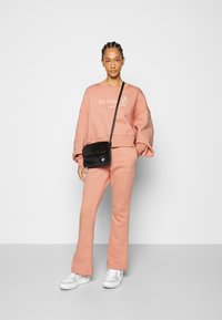 Sweater oversized rosa claro com o texto "Seja gentil", acompanhado de calças de moletão ajustadas combinantes. Bolsa preta de tiracolo e sapatilhas brancas.