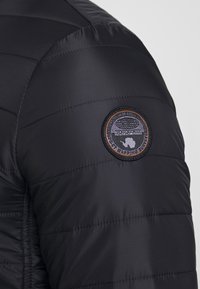 Schwarze Pufferjacke mit horizontalen Nähten, ausgestattet mit einem runden Patch in Orange und Grau auf dem Ärmel, der ein strukturiertes, glänzendes Material zeigt.