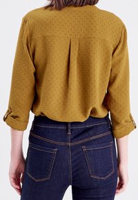 Blouse jaune moutarde, texturée, avec des manches retroussées et un pli au dos, associée à un jean en denim bleu foncé avec des coutures contrastées et des poches.