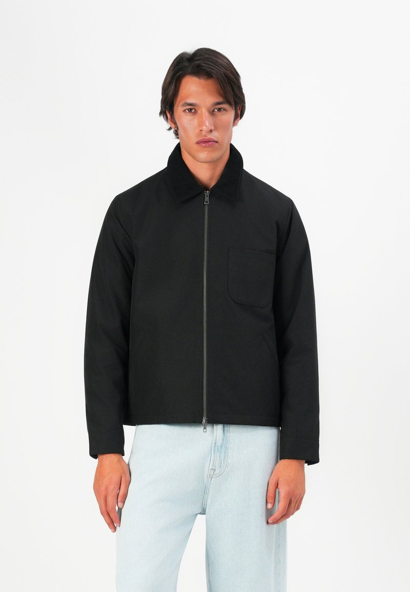 Black zip-up jacket with a collared neckline, front pocket, and a structured fit. Worn over light blue jeans. Smooth fabric texture.