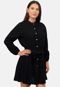 Abito nero con bottoni, realizzato in un tessuto testurizzato, maniche lunghe, cintura da annodare in vita e colletto arrotondato.
