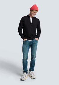 Schwarze Zip-Jacke mit Stehkragen, getragen über einem weißen Hemd. Blaue Jeans und weiße Sneaker. Rote Strickmütze als Akzent.