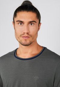 Grå t-shirt med rund hals og navyblå kant, med et lille broderet logo på brystet og en glat, let stoftekstur.