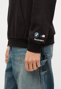 Άτομο που φοράει μαύρο φούτερ BMW Motorsport και μπλε τζιν με το χέρι στην τσέπη, σε λευκό φόντο.