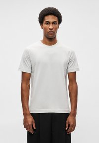 Svetlo šedé bavlnené tričko s okrúhlym výstrihom, krátkymi rukávmi a nenápadným bielym logom vytlačeným na hrudi. Jednoduchý dizajn, voľný strih.