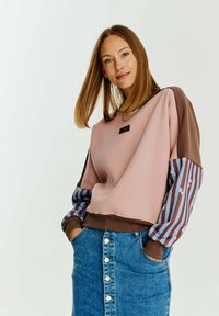 Mikina s ružovým telom a hnedými akcentmi. Pruhované modré a hnedé rukávy s kvetinovými detailmi. V kombinácii s modrou džínsovou sukňou s gombíkmi.
