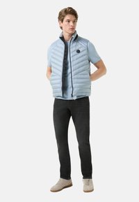 Light blue puffer vest with a high collar, horizontal quilted design, black zipper, worn over a light blue t-shirt and dark jeans, beige sneakers.