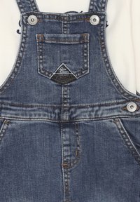 Salopette in denim con spalline regolabili, dotate di tasca frontale e un patch a forma triangolare. Completate da accessori in metallo e cuciture a contrasto.