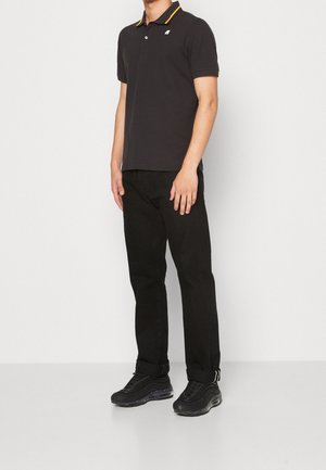 Mężczyzna ubrany w czarną koszulkę polo z żółtymi akcentami, czarne dżinsy z podwiniętymi nogawkami i czarne sneakersy, stojący na białym tle.